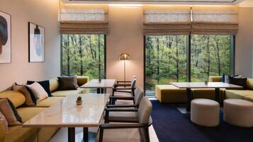 A modern hotel room with a sofa, tables, and a view of a forest through large windows.