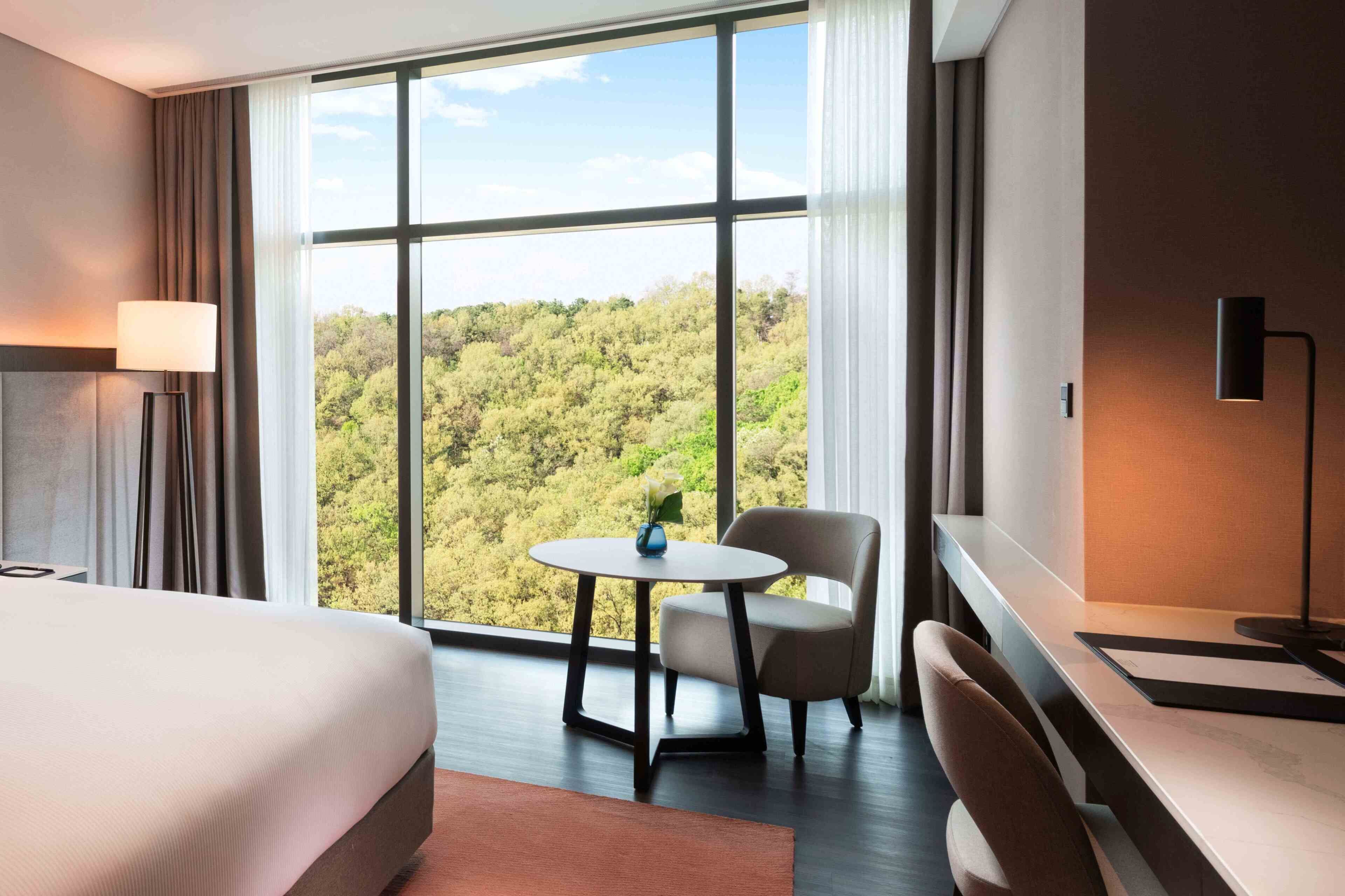 Habitación de hotel con un ventanal grande, una cama, un escritorio, una silla y una lámpara.