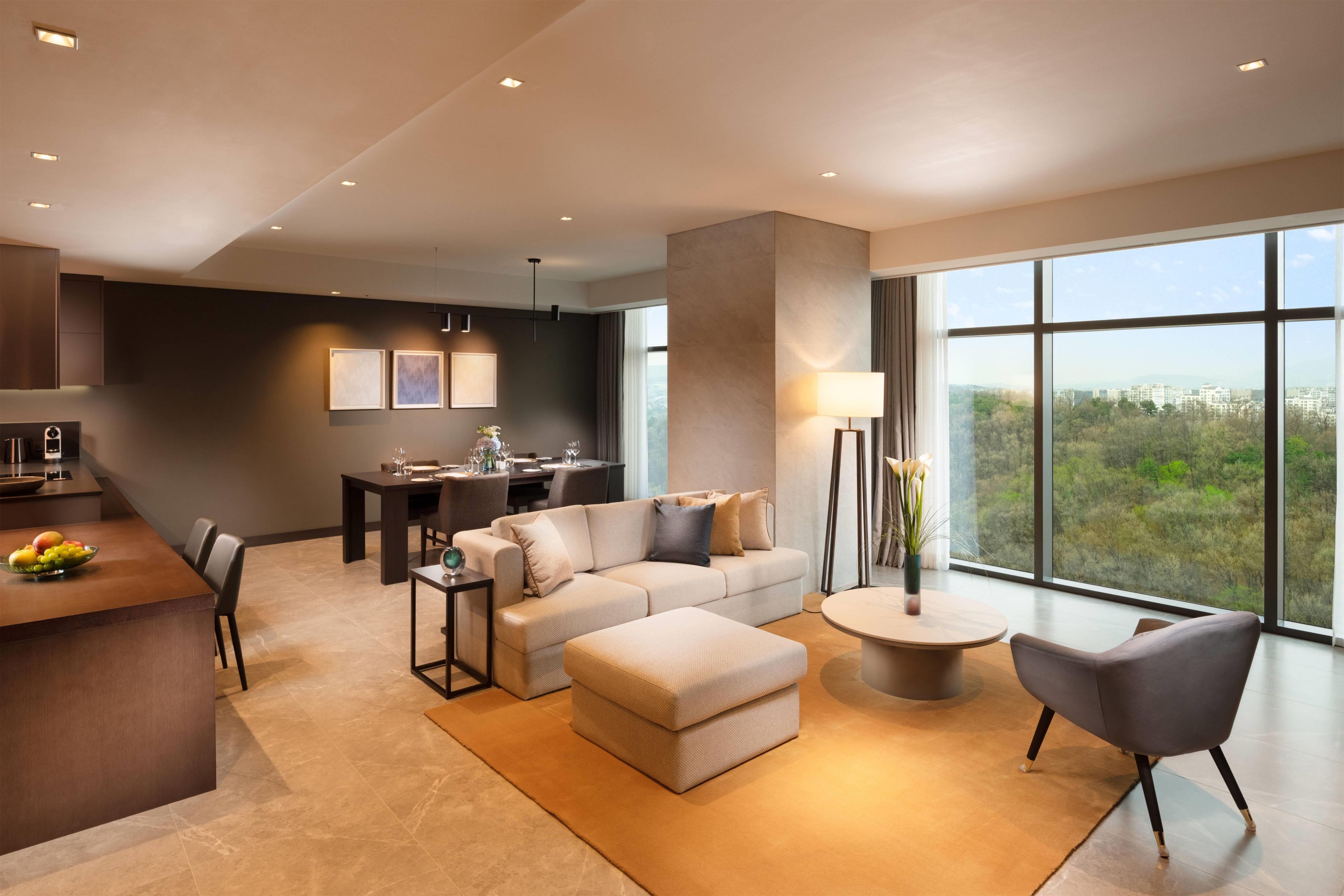 A modern living room with a sofa, ottoman, and coffee table, a dining area with a table and chairs, and a kitchen area with a fruit bowl.