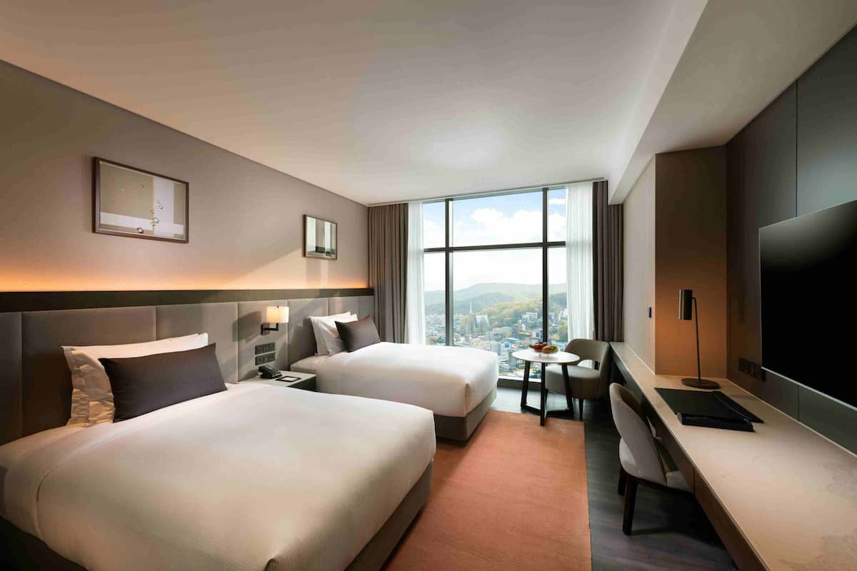 Habitación de hotel con dos camas, un escritorio, una silla, un televisor y un ventanal grande con vistas a la ciudad.
