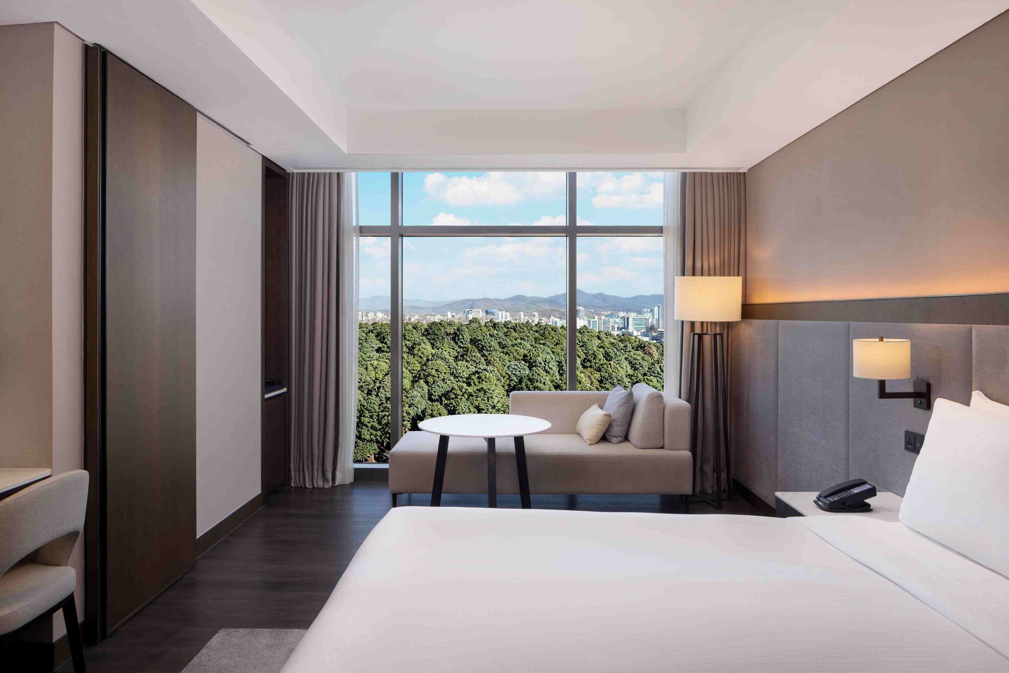 Habitación de hotel con cama, sofá, mesita, teléfono y vistas a la ciudad y montañas a través de amplias ventanas.