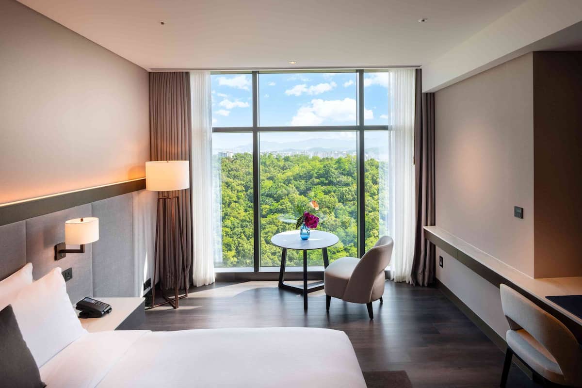 Habitación de hotel con un ventanal grande, una cama, un escritorio, una silla y una pequeña mesa con un florero.