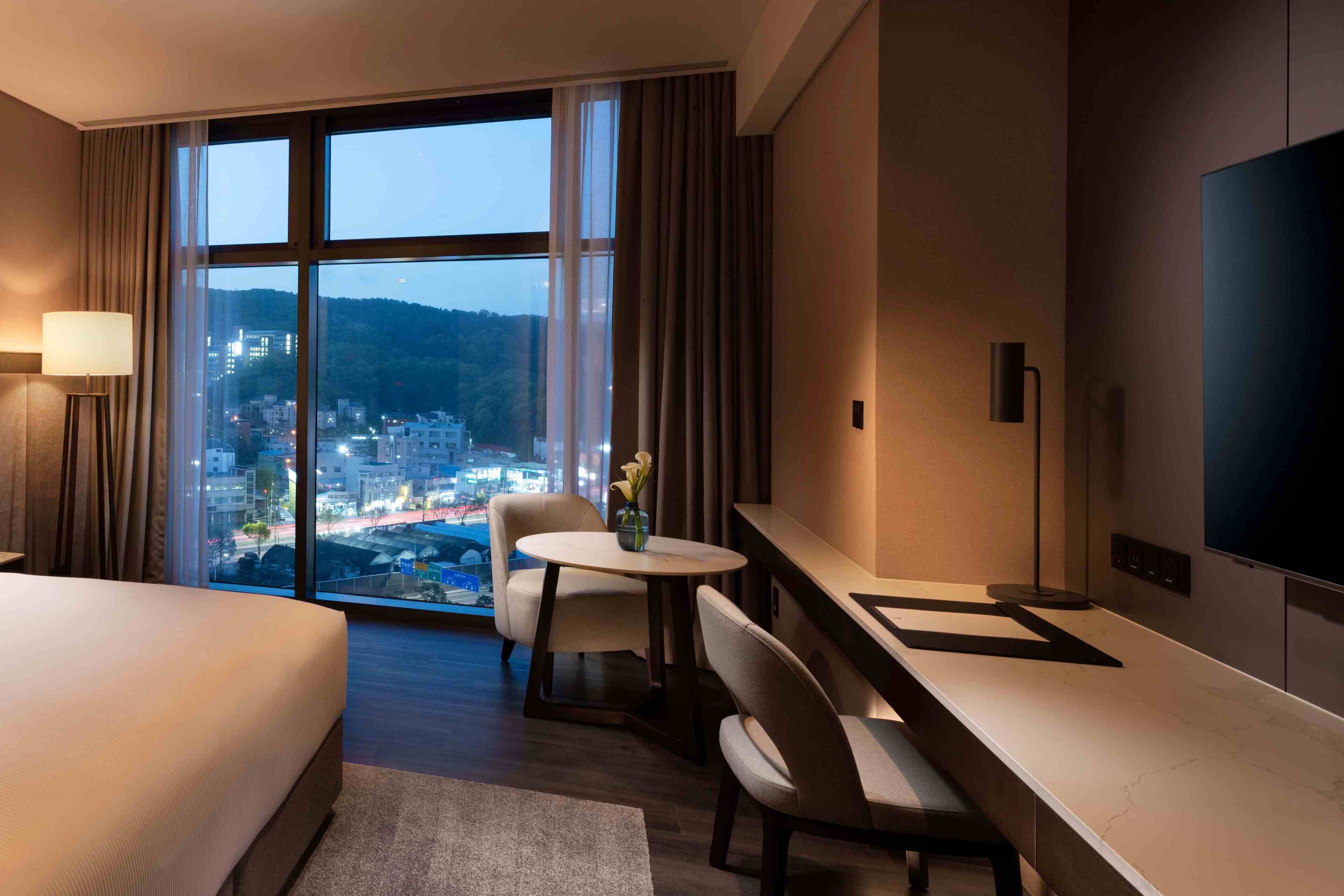 A hotel room with a bed, a desk, a chair, a TV, and a large window with a city view.