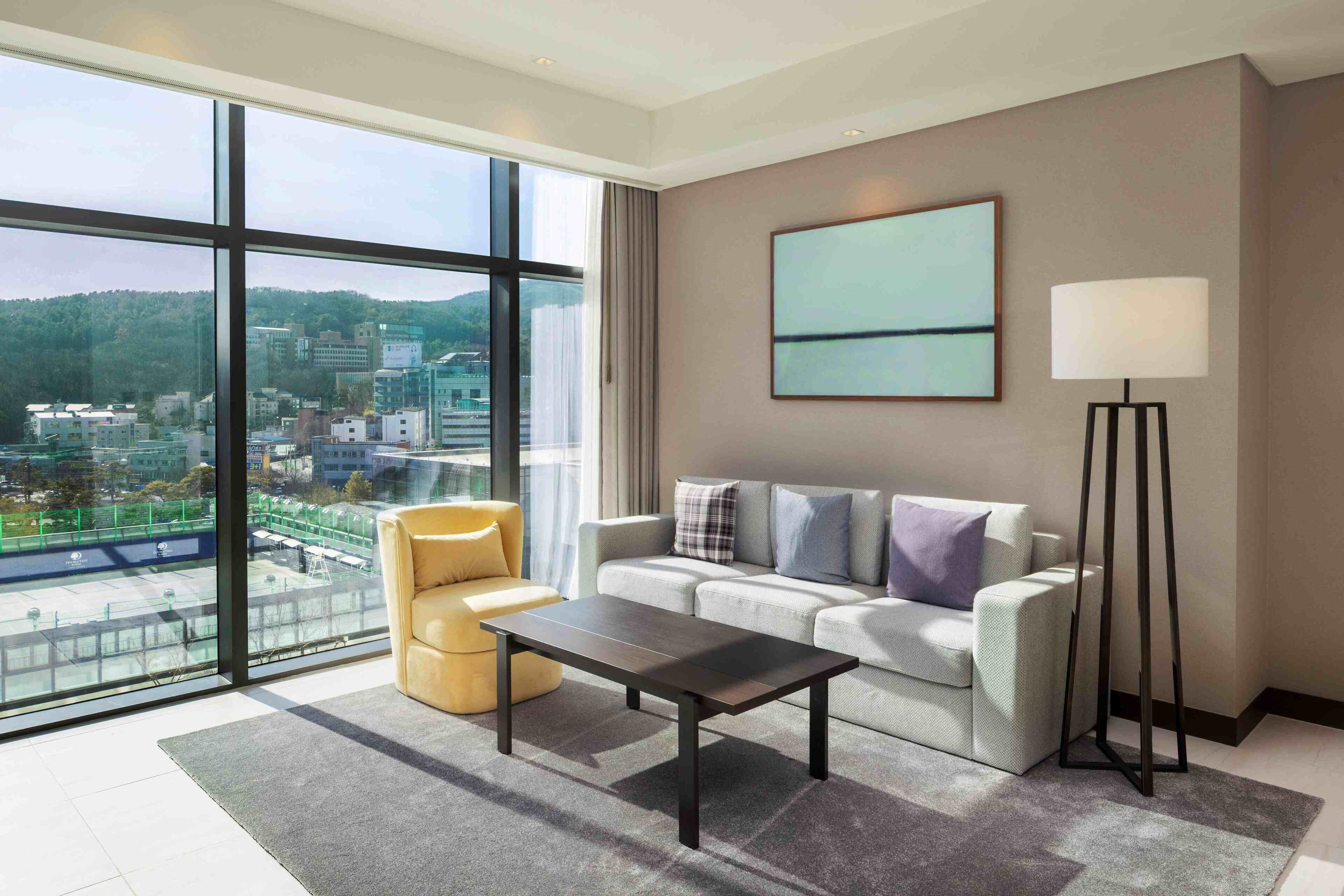 A modern living room with a sofa, a coffee table, a floor lamp, and a large window offering a city view.