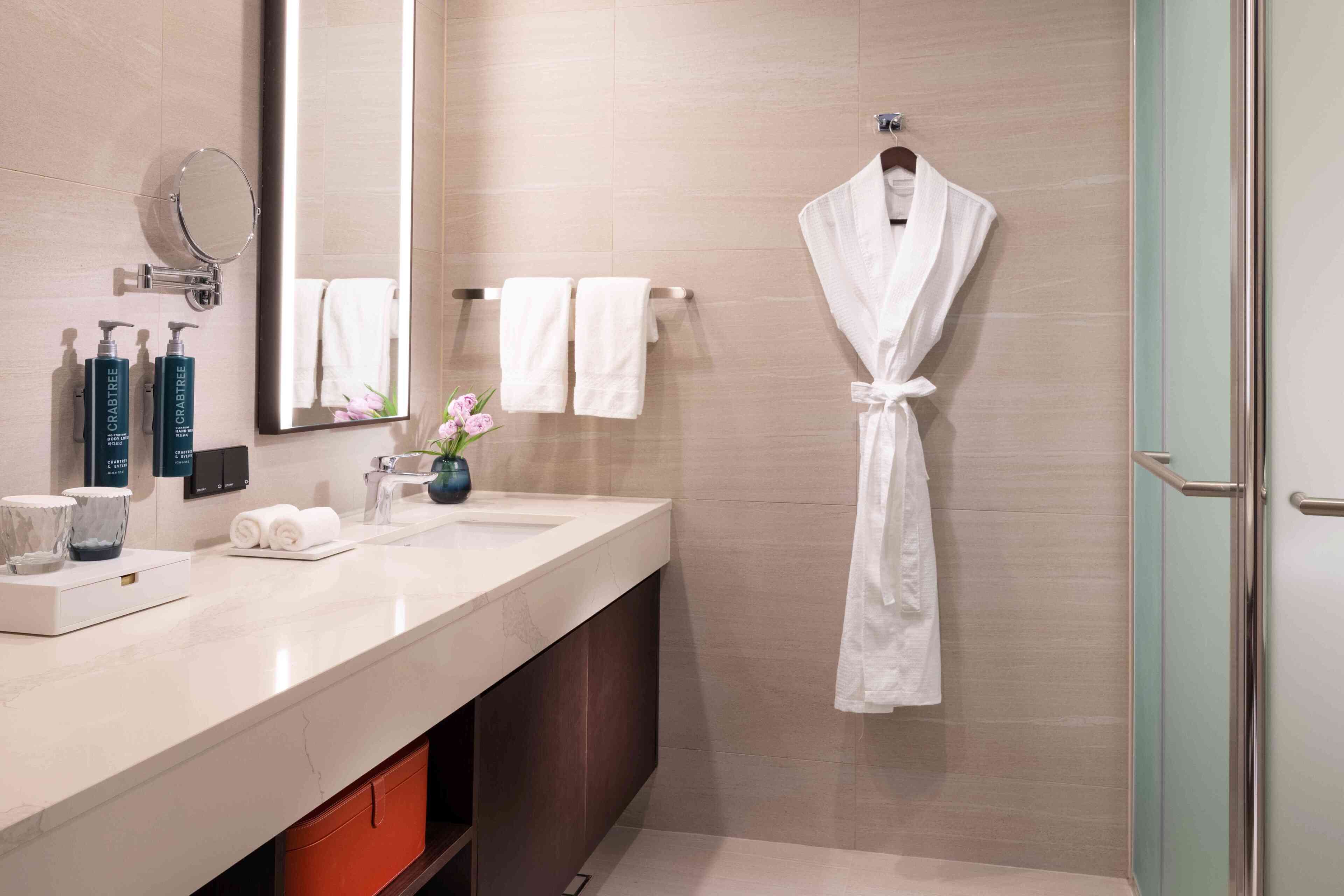 Une salle de bain moderne avec un grand miroir, deux vasques, un peignoir suspendu à un porte-peignoir et une porte de douche en verre.