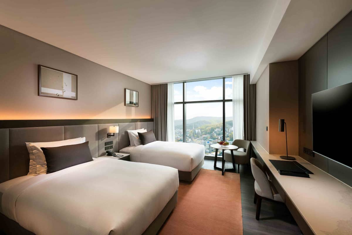 Habitación de hotel con dos camas, un escritorio, una silla, un televisor y un ventanal con vista a la ciudad.