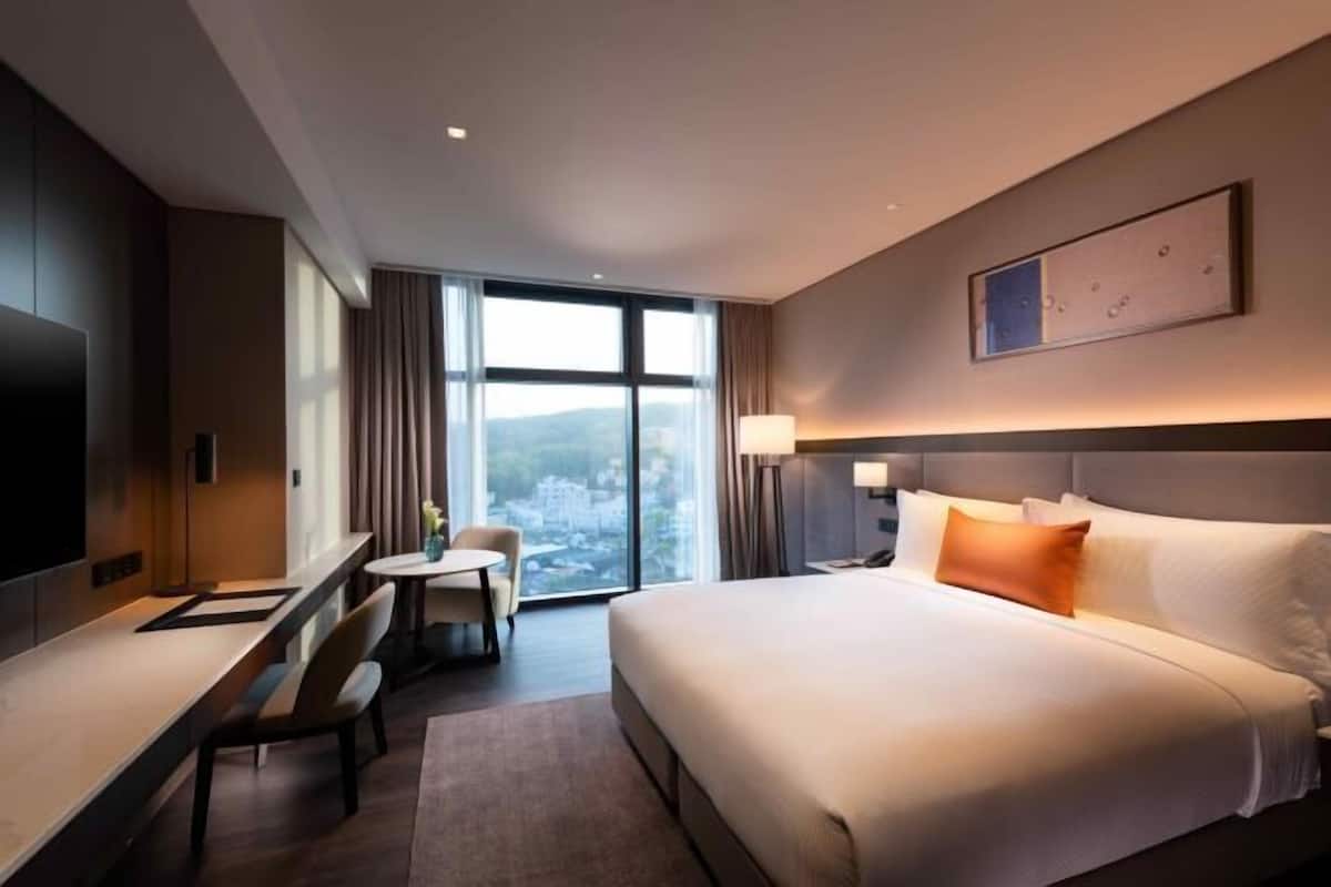 Habitación de hotel con cama, escritorio, silla, lámpara y un gran ventanal con vistas.