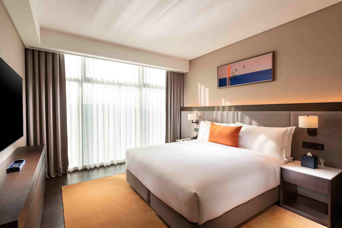 Habitación de hotel con una cama grande, un televisor de pantalla plana, una mesita de noche y un ventanal con cortinas.