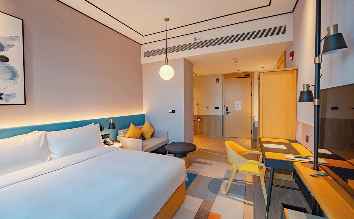 Ein modernes Hotelzimmer mit einem großen Bett, einer Couch, einem Schreibtisch und einem Sessel. Ein Bild hängt an der Wand und eine Tür führt in einen anderen Raum.