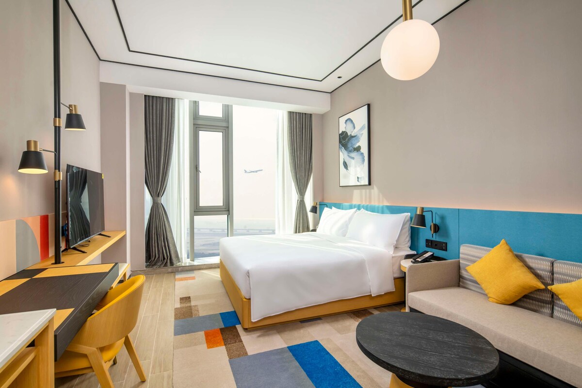 A hotel room with a bed, a sofa, a desk, a TV, and a large window.