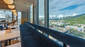 A modern restaurant with a long bench seating area, large windows offering a city view, and contemporary lighting fixtures. - Hilton Garden Inn Shenzhen Airport (Shenzhen)
