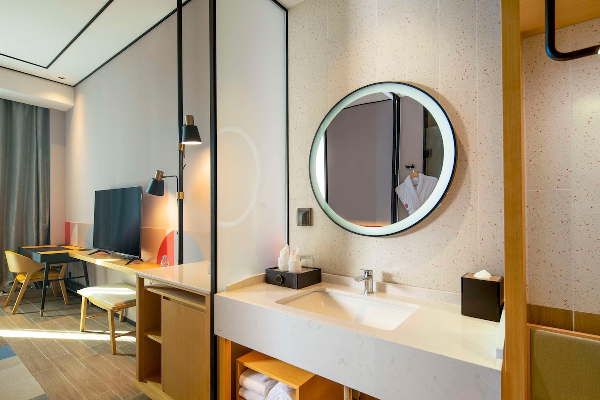 Une salle de bain d’hôtel moderne avec un miroir rond, un lavabo et une coiffeuse.