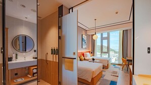 A modern hotel room with a large bed, a sitting area, a bathroom with a mirror and sink, and a view of the city. - Hilton Garden Inn Shenzhen Airport (Shenzhen)