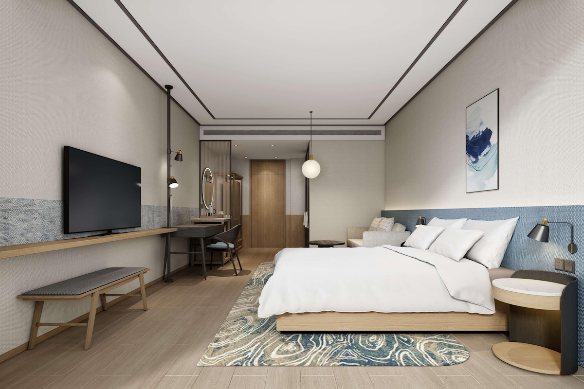 A modern hotel room with a bed, desk, chair, TV, and a bathroom mirror.