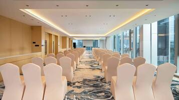 A hotel conference room with rows of white chairs, a large screen at the front, and a view of the city through large windows.