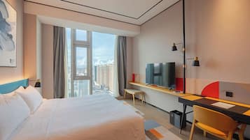 A hotel room with a large bed, a desk with a chair, a TV, and a view of the city.