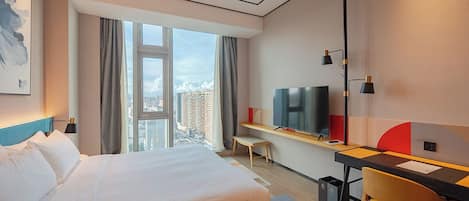 A hotel room with a large bed, a desk with a chair, a TV, and a view of the city.