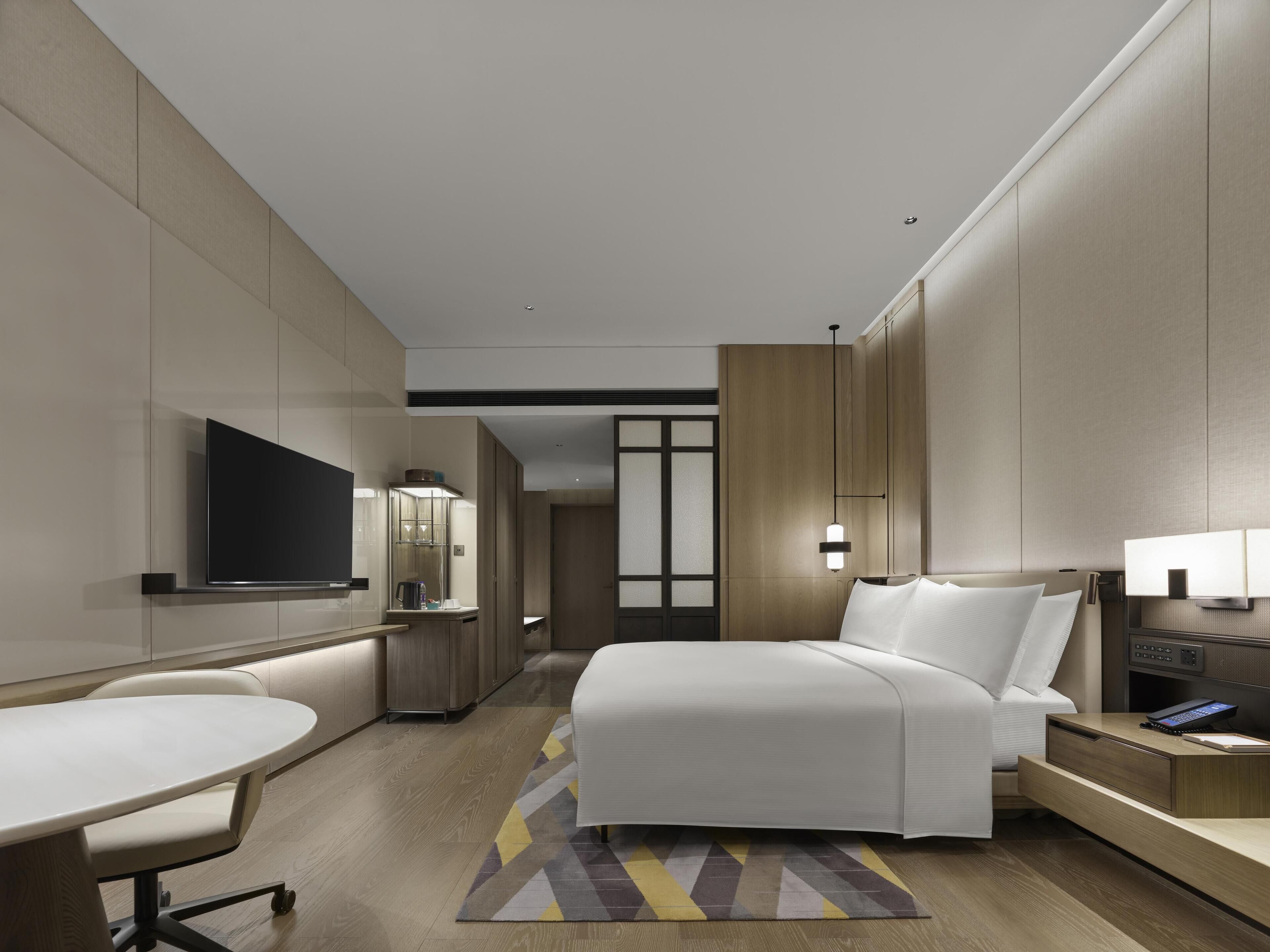 A modern hotel room with a bed, bedside table, lamp, TV, and a desk with chair.