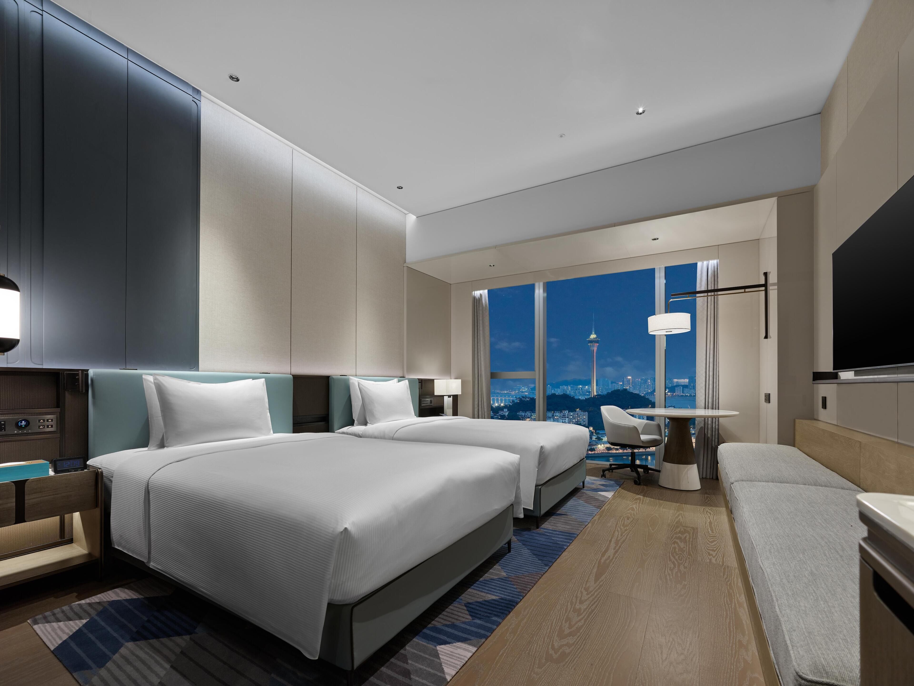 A modern hotel room with two beds, a desk, a chair, and a large window offering a city view.