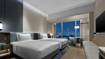 A modern hotel room with two beds, a desk, a chair, and a large window offering a city view.