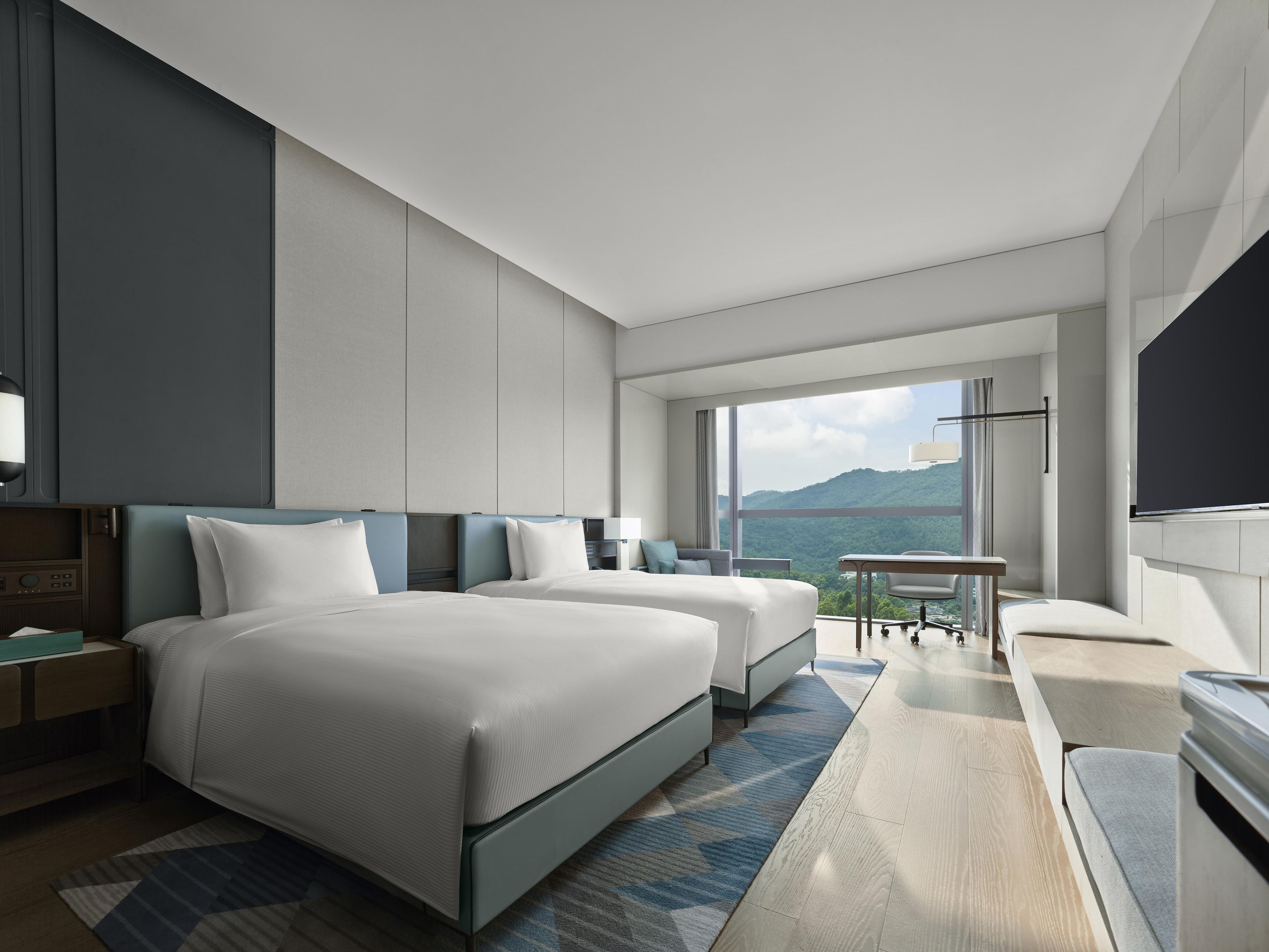 Ein modernes Hotelzimmer mit zwei Betten, einem Schreibtisch und einem großen Fenster mit Blick auf die Berge.