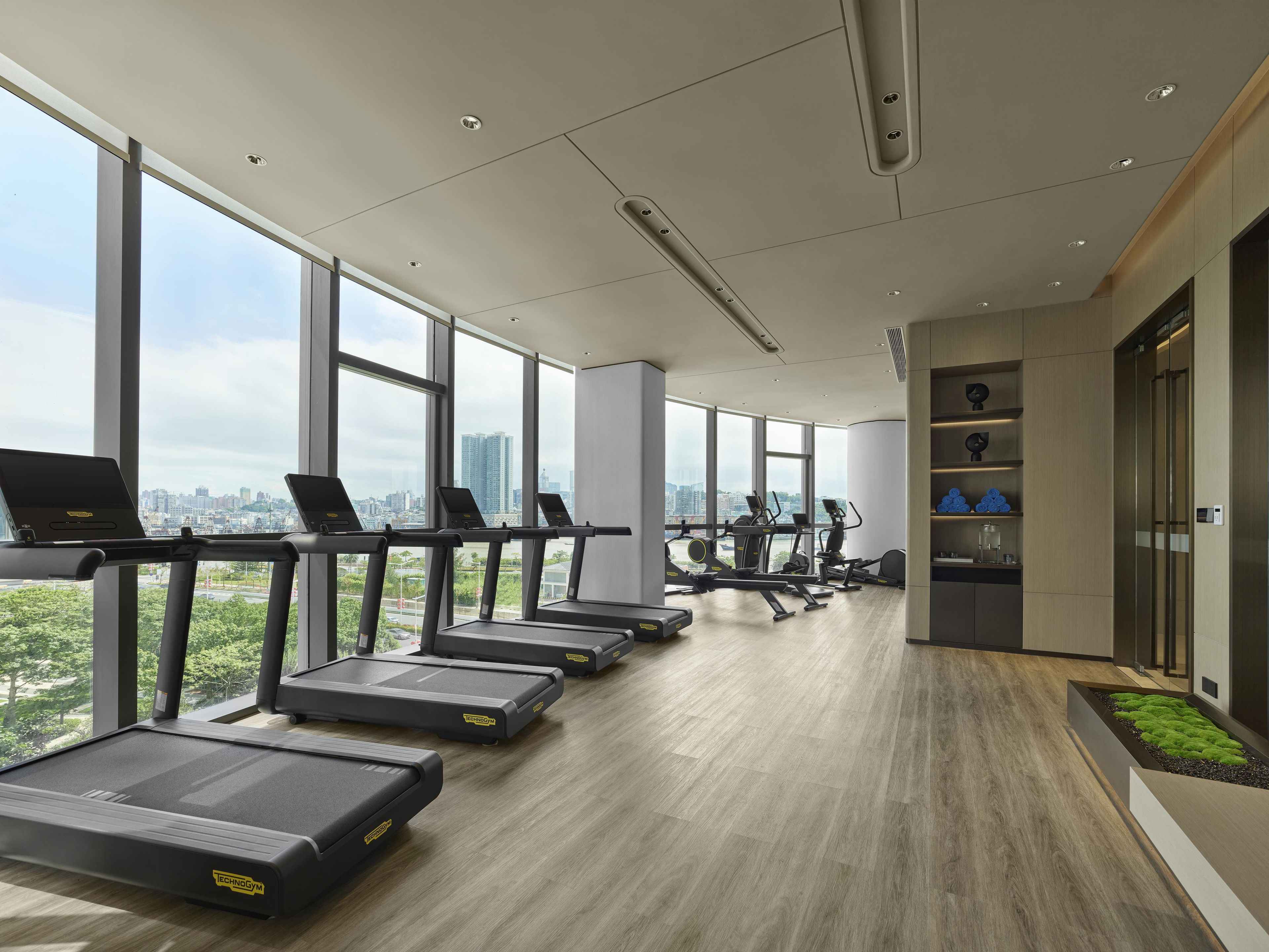 A gym with treadmills, exercise bikes, and a city view through large windows.