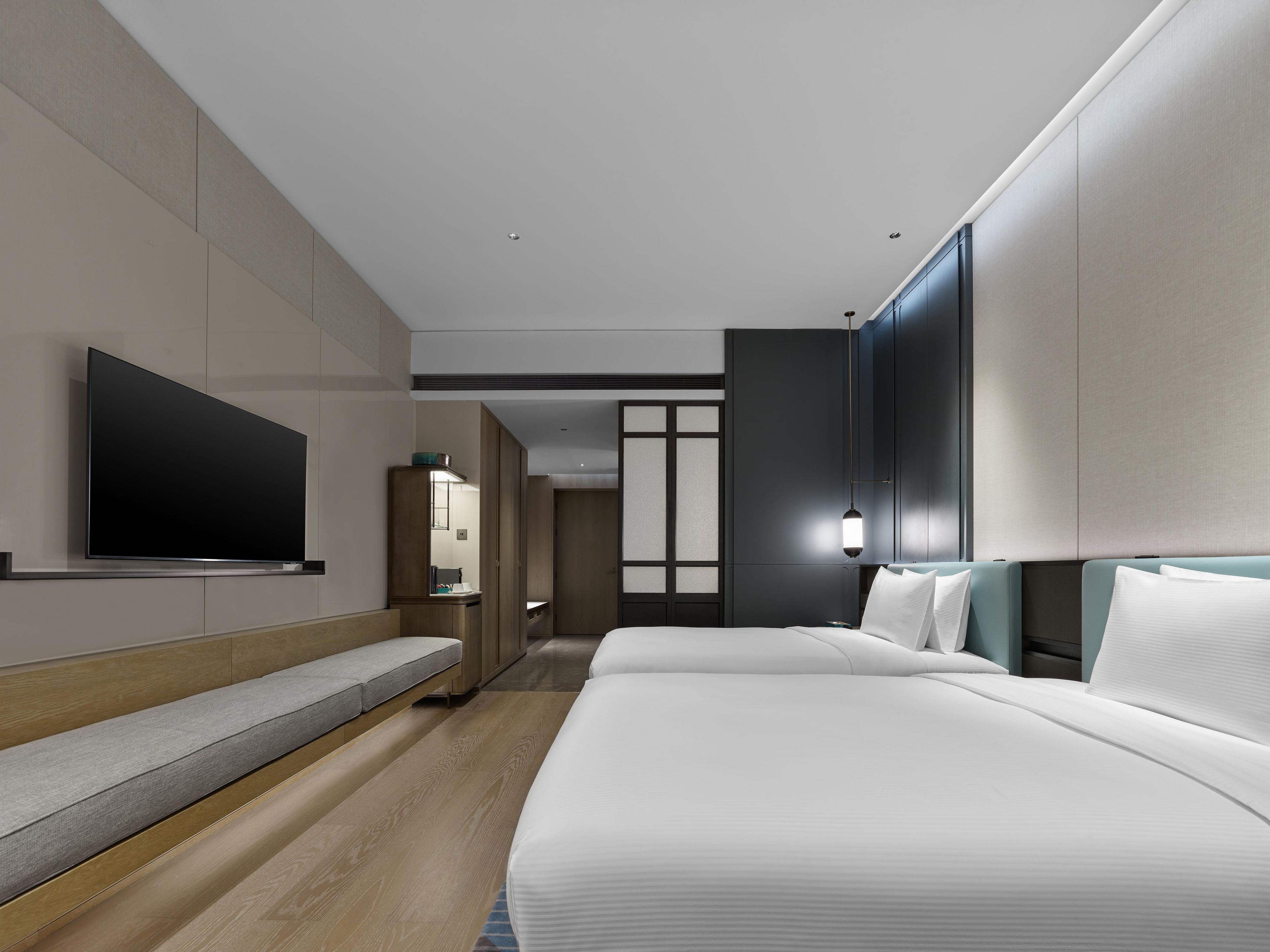 A modern hotel room with a large bed, a flat-screen TV mounted on the wall, a seating area with a bench, and a walk-in closet.