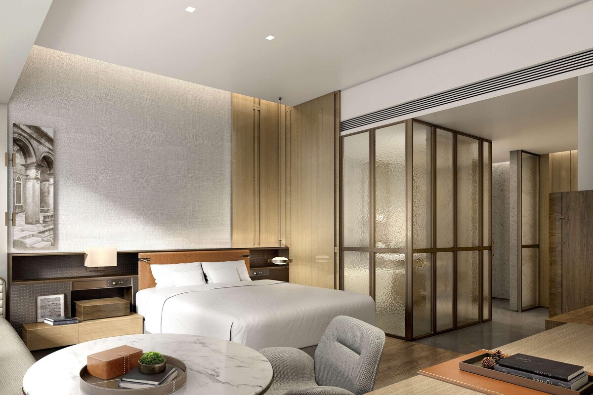 A modern hotel room with a large bed, a sofa, a small table with a plant, and a view of the city.