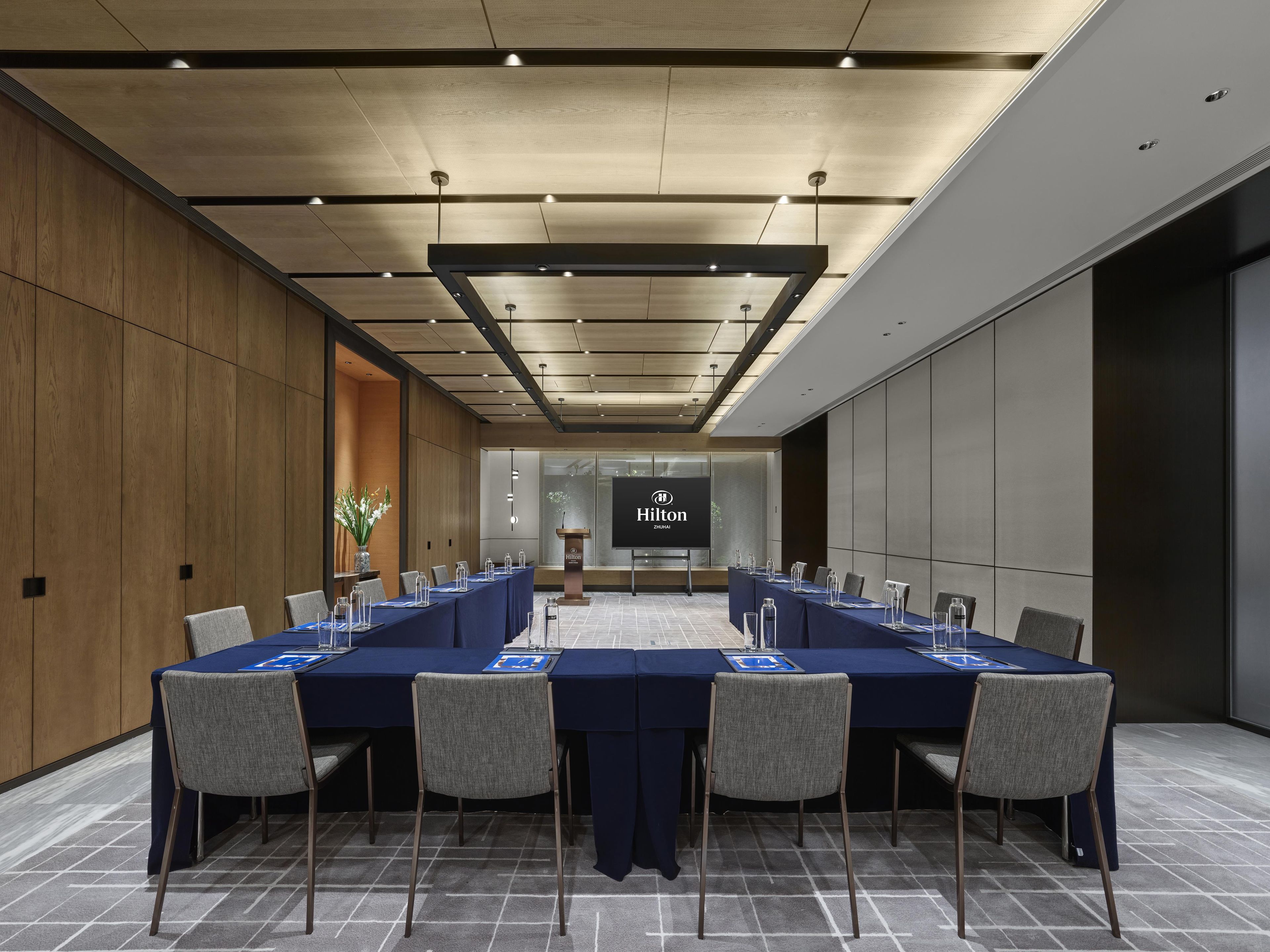 Un salón de conferencias con una larga mesa, sillas, un atril y una pantalla que muestra el logotipo de Hilton.