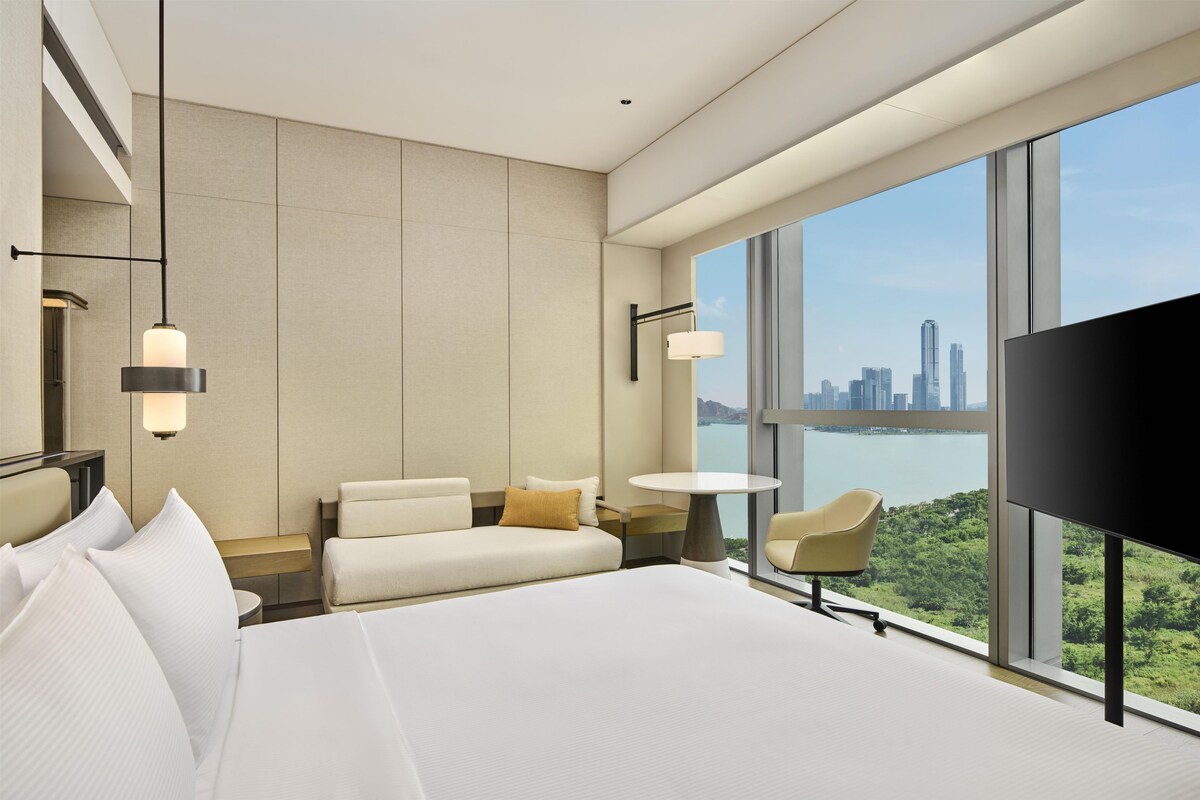 Una habitación de hotel moderna con una cama grande, una zona de estar con una mesa y vistas a la ciudad a través de amplios ventanales.