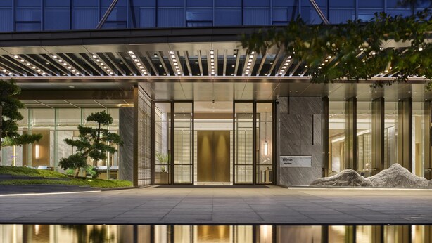 Una entrada de hotel moderna con fachada de vidrio, un estanque reflectante e iluminación exterior.