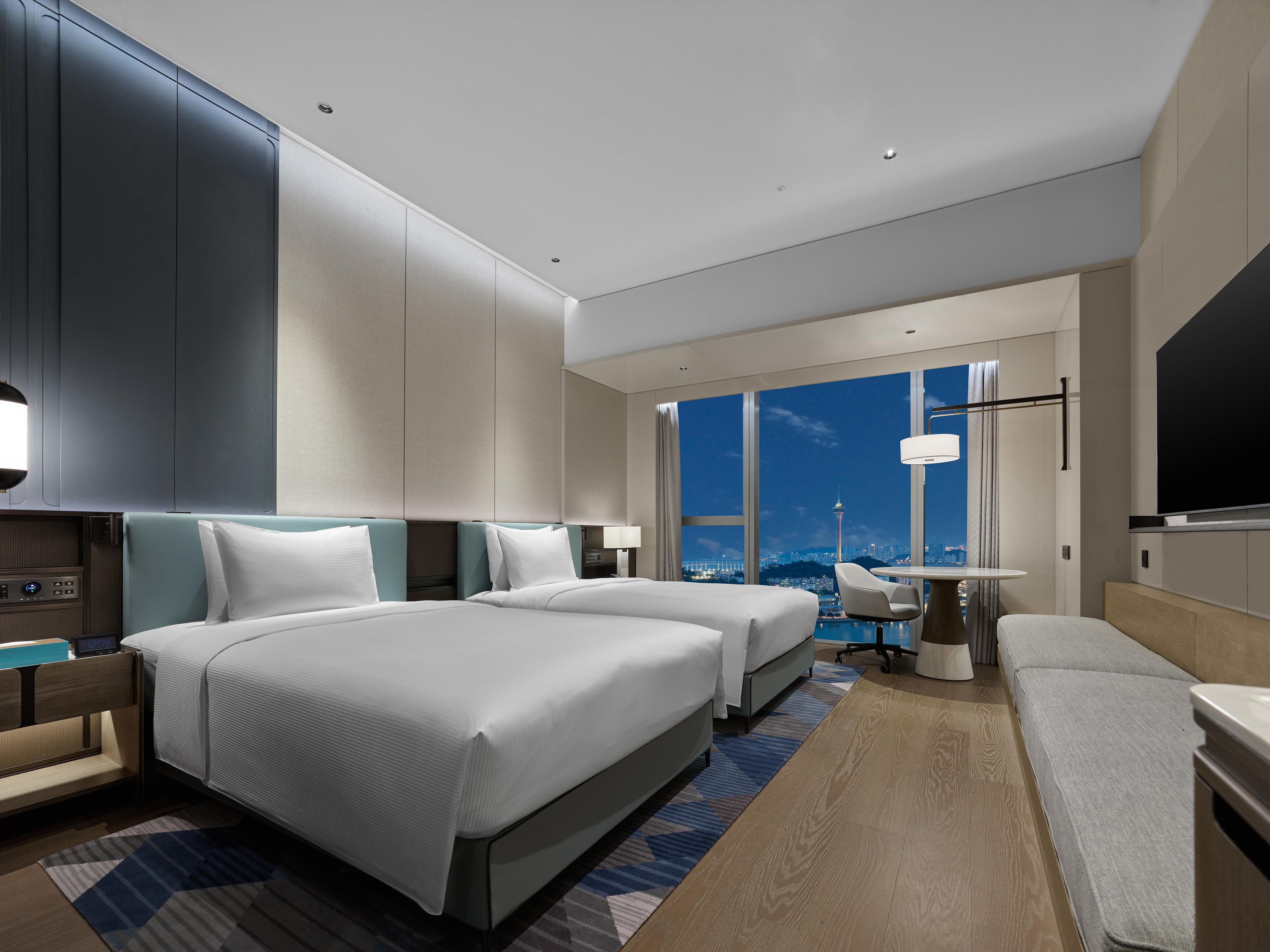 A modern hotel room with two beds, a desk, a chair, a TV, and a large window offering a city view.