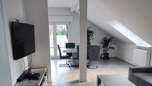 A modern living room with a flat-screen TV, a coffee table, and a sofa. There is a dining area with a table and chairs in the background.