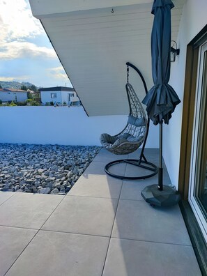 A patio with a hanging chair and umbrella, a tiled floor, and a view of houses in the background. - Ferienwohnung Schwäbische Albkante (Dürnau)