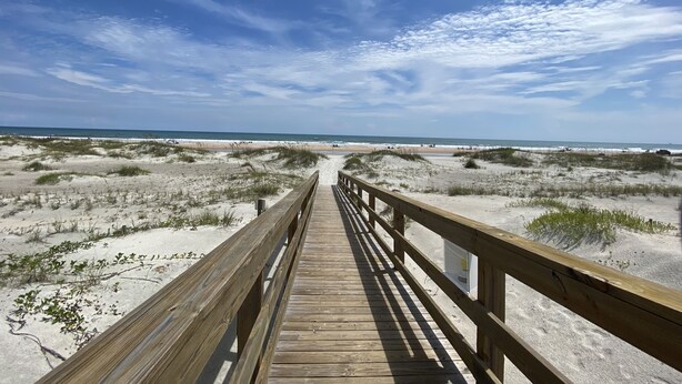 A wooden boardwalk leading to a beach with dunes and a clear sky.