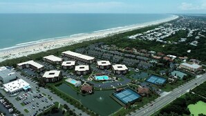 An aerial view of a coastal resort with multiple buildings, a swimming pool, tennis courts, and a beach in the background.