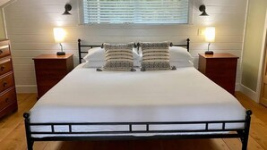 Een net opgemaakt bed in een kamer met houten vloeren, witte wanden en een schuin dak. Er staan twee nachtkastjes met lampen, een dressoir en een raam met zonwering.