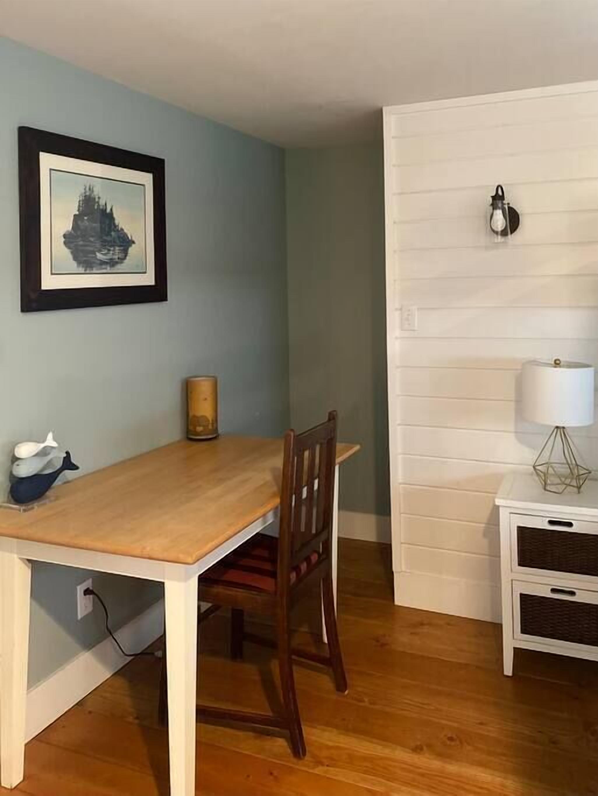 A dining area with a wooden table and chairs, a framed picture on the wall, a lamp on a side table, and a white cabinet with drawers.