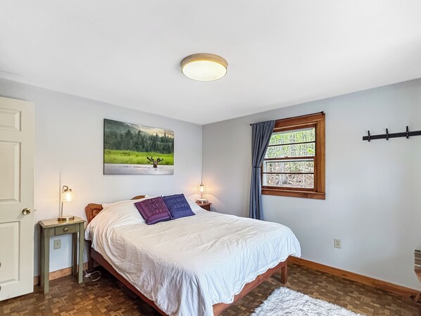 Un dormitorio con cama, mesita de noche, lámpara, ventana con cortinas, un cuadro en la pared y una luz en el techo.