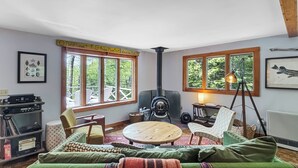 Ett mysigt vardagsrum med en soffa, fåtöljer, ett runt träbord, en öppen spis och stora fönster med utsikt över grönområden.