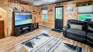 Ein Holzblockhaus-Interieur mit einer Holztreppe, einem Fernseher, der eine Strandansicht zeigt, einem schwarzen Ledersofa und einem gemusterten Teppich.