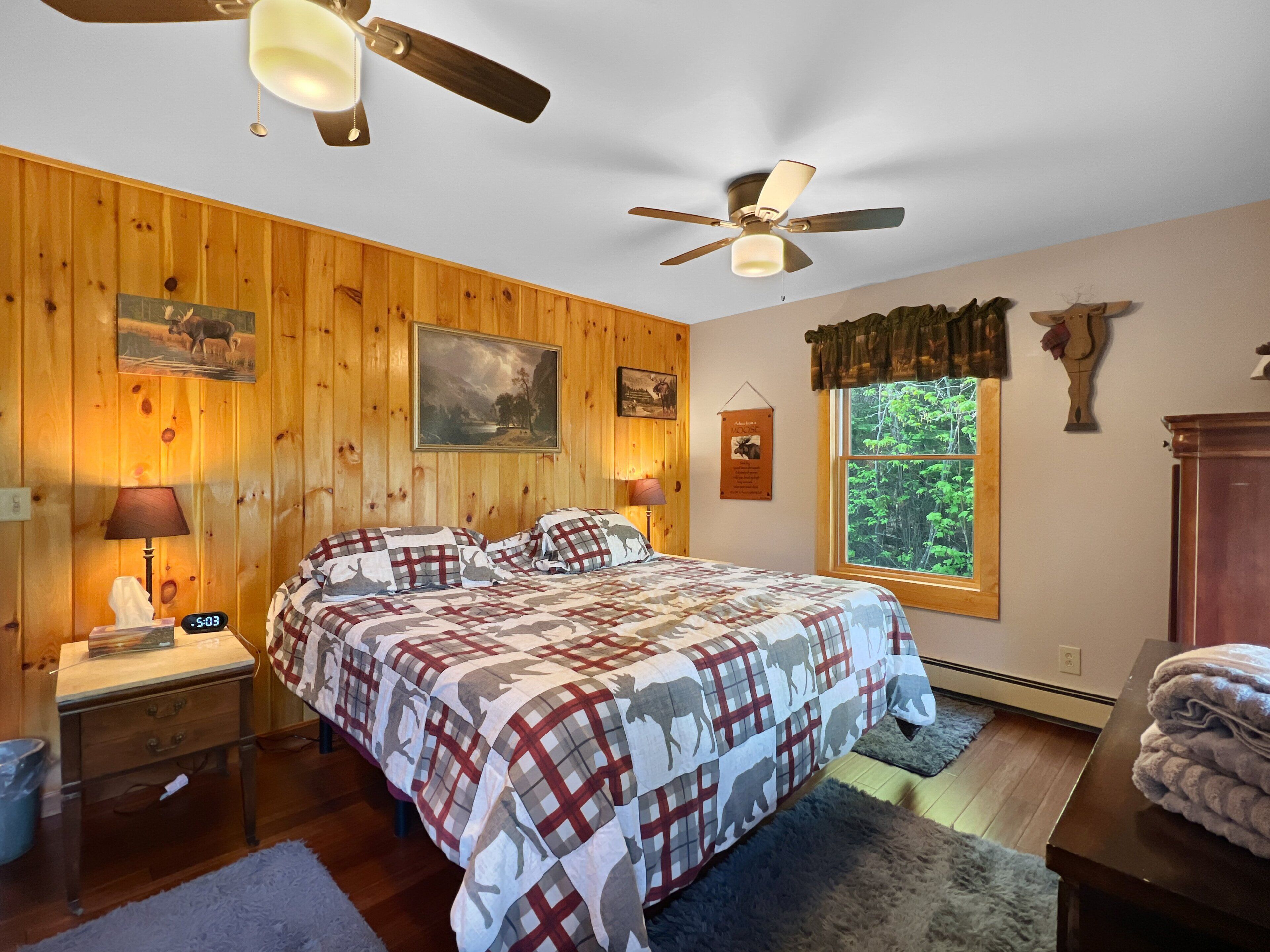 A bedroom with a wooden wall, a bed with a plaid bedspread, two bedside lamps, a ceiling fan, and a window with a view of trees.