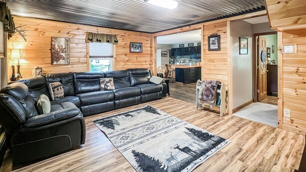 Ein rustikales Blockhaus-Interieur mit einer Ledersofa, einem gemusterten Teppich, Holzwänden und einem durch eine offene Tür einsehbaren Küchenbereich.