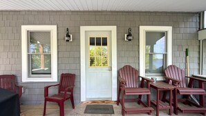 Eine überdachte Veranda mit einer grauen Außenwand, einer weißen Tür und einer Gruppe Adirondack-Stühle.
