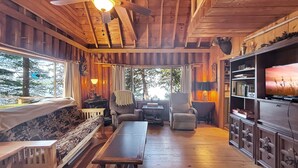 A cozy cabin interior with a wooden ceiling, a ceiling fan, a television, a fireplace, and a large window overlooking a forest. - Horizons West Hideaway (Waterfront on Moosehead Lake) (Greenville)