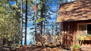 A cabin with a wooden deck, a thatched roof, and a view of a lake surrounded by tall trees. - Horizons West Hideaway (Waterfront on Moosehead Lake) (Greenville)