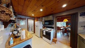 A compact kitchen with wooden cabinets, a microwave, a stove, and a dining area with chairs. - Horizons West Hideaway (Waterfront on Moosehead Lake) (Greenville)