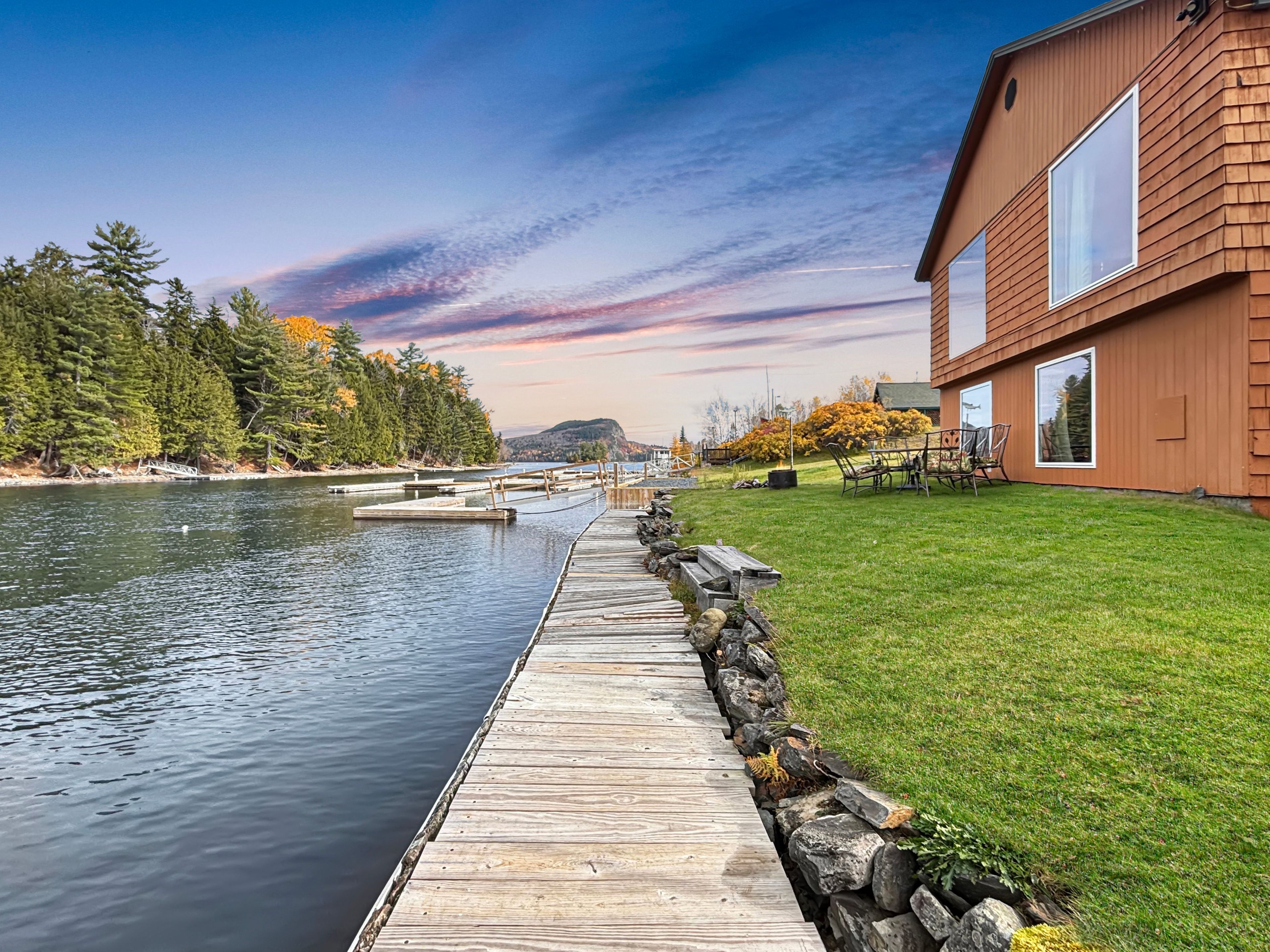 A wooden dock extending into a calm body of water, with a house on the right featuring large windows and a grassy yard.