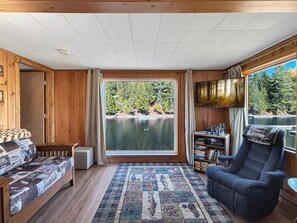 A cabin with a large window overlooking a lake, a sofa, a chair, and a bookshelf.