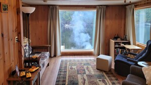 Ein gemütliches Blockhauss-Interieur mit großem Fenster, Holzwänden, einer Couch, einem Lesesessel und einem kleinen Tisch mit Büchern und einer Lampe.